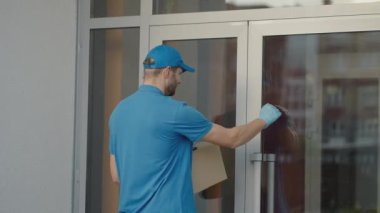Caucasian young pretty man, delivery worker in blue cap walking the street and carrying carton box while using smartphone, looking for route. Male courier with parcel tapping and texting on phone.
