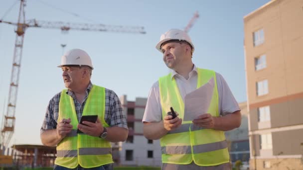 建築現場に立つ2人の建築家の肖像。ヘルメットとベストの建設中の建物の背景に立って図面を持つ2人の建築家 — ストック動画