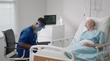 Koruyucu maskeli siyahi bir erkek doktor hastane yatağında yatan yaşlı beyaz bir adamla hastalarıyla konuşuyor.