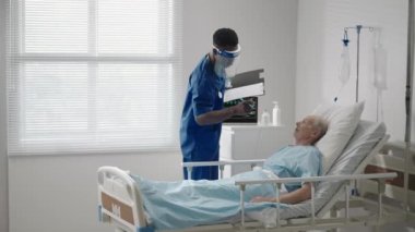 ECG makinesine bağlı olarak hastanede yatan yaşlı bir erkek hasta koruyucu maskeli siyahi bir doktorla konuşuyor.