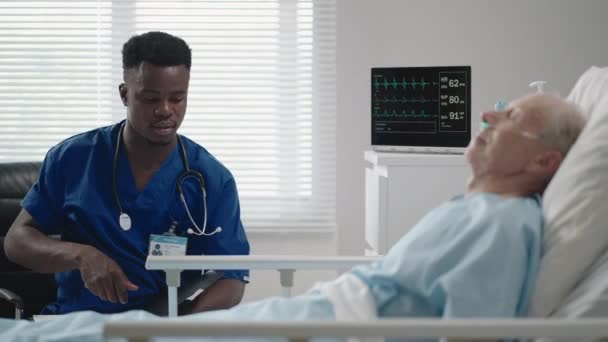 Un médico cardiólogo negro está hablando con un paciente de 60-70 años de edad, acostado en una cama en un hospital. Un neurólogo está hablando con un paciente. El paciente está conectado a una máscara de oxígeno y un dispositivo de ECG — Vídeo de stock