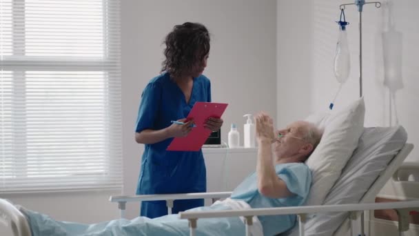 En mörkhyad kvinnlig läkare pratar med en gammal manlig patient som ligger på en sjukhussäng och diskuterar rehabilitering efter tillfrisknandet.. — Stockvideo
