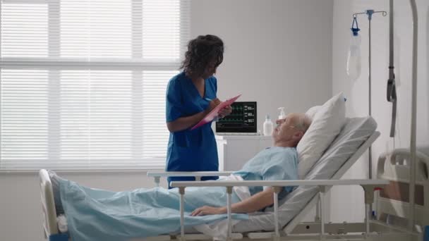 Ein älterer weißer Mann im Alter von 60-70 Jahren liegt in einem Krankenhausbett und spricht mit einer schwarzen Ärztin. Diskussion über die Behandlung mit einem kardiologen Neurologen. Rehabilitationsplan nach Genesung — Stockvideo
