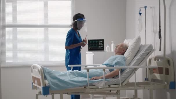 En svart kvinnlig kardiolog läkare pratar med en gammal man patient som ligger på en sjukhussäng och diskuterar rehabilitering efter tillfrisknandet. en infektionssjukdomsneurolog diskuterar en behandlingsstrategi — Stockvideo