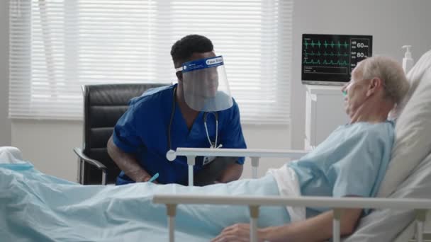 Koruyucu maskeli siyahi bir erkek doktor hastane yatağında yatan yaşlı beyaz bir adamla hastalarıyla konuşuyor. — Stok video