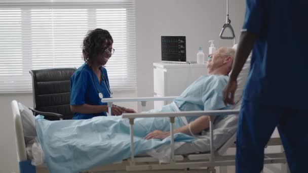 Африканская команда молодых врачей консультирует больного ковидом-19 в постели в больнице. Больной лежит на больничной койке и разговаривает с хирургом в халате и защитной маске. врач осматривает пожилого пациента — стоковое видео