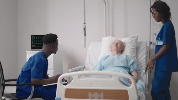 Afrikalı etnik grup doktorlarından oluşan bir ekip yaşlı bir erkek ve kadınla konuşuyor. Bir doktor ve bir hemşire hastane koğuşunda yatalak bir hastayla konuşuyorlar. — Stok video