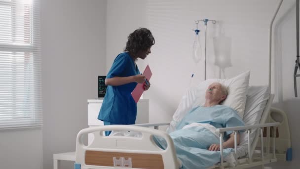 En svart kvinnlig kirurg pratar med en äldre manlig patient som ligger på en sjukhussäng. En kvinnlig kardiolog infektionssjukdomsspecialist talar med en patient på kliniken — Stockvideo