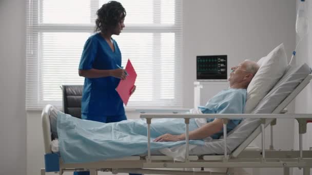 Een vrouwelijke dokter met een donkere huid praat met een oude mannelijke patiënt liggend op een ziekenhuisbed over rehabilitatie na herstel.. — Stockvideo