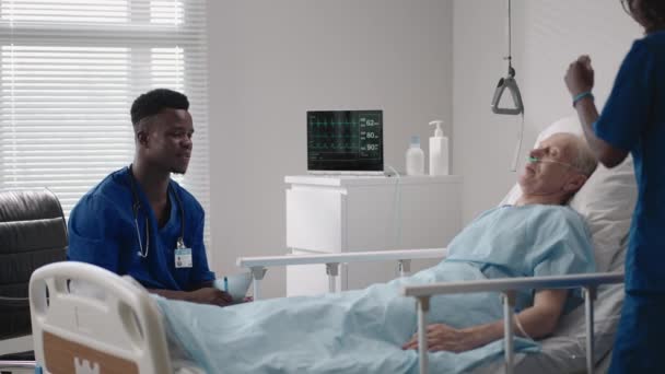 Yaşlı bir erkek hastanın yatağının yanında Afrikalı etnik tıp ekibine bakmak. Bakım görevlileri yaşlı bir erkek hastane hastasının hayati belirtilerini izliyor. — Stok video