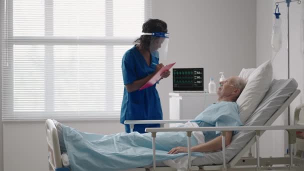 En svart kvinnlig kirurg pratar med en äldre manlig patient som ligger på en sjukhussäng. En kvinnlig kardiolog infektionssjukdomsspecialist talar med en patient på kliniken — Stockvideo