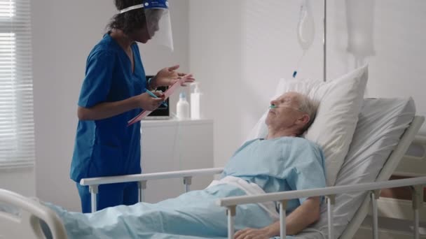 A black female surgeon is talking to an elderly male patient lying on a hospital bed. A female cardiologist infectious disease specialist is talking to a patient in the clinic — Stock Video