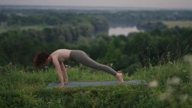 Ağır çekim: Eşofmanlı genç kadın dağlarda yoga pozisyonu alıyor. Kamera bir şeyler yaratıyor. Dışarıda yoga yapan bir kadın. Yoga yapan bir kadın..