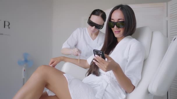 Podczas laserowej sesji usuwania włosów, kobieta w salonie piękności pisze wiadomość w telefonie komórkowym. Depilacja ręczna depilacja zdjęcie depilacja i kobieta pisze wiadomość w smartfonie — Wideo stockowe