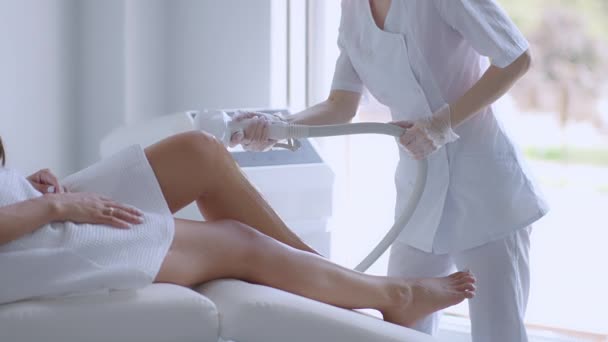 Женщина лежит на диване для лазерной процедуры по удалению волос. Аппаратное удаление волос на ногах в салоне красоты. Лазерная эпиляция женских ног. Руки косметолога делают эпиляцию — стоковое видео