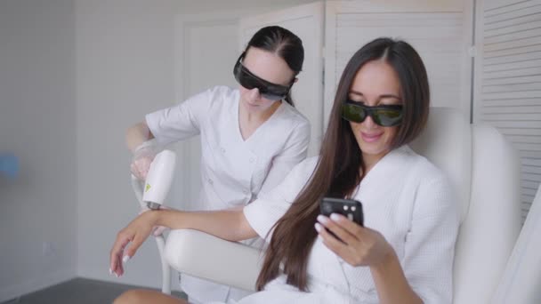 レーザー脱毛セッションでは、美容室の女性が携帯電話にメッセージを書き込みます。手の毛の除去写真の毛の除去と女性はスマートフォンでメッセージを書きます — ストック動画