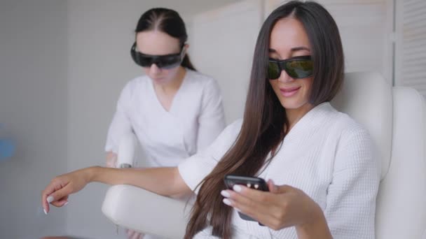 Under en laser hårborttagning, en kvinna i en skönhetssalong skriver ett meddelande i sin mobiltelefon. Hand hårborttagning foto hårborttagning och en kvinna skriver ett meddelande i en smartphone — Stockvideo
