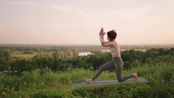A young woman does yoga exercises standing on the top of a forest mountain against the background of a river and a forest landscape. Balance of body and spirit, relaxation and meditation — Stock Video