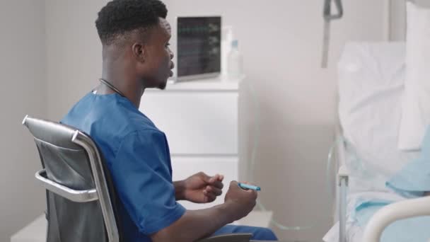 アフリカの男性医師が酸素マスクで病院のベッドに横たわっている患者をインタビューします。病院のベッドに寝そべっている黒人女性は医者に症状を説明する — ストック動画
