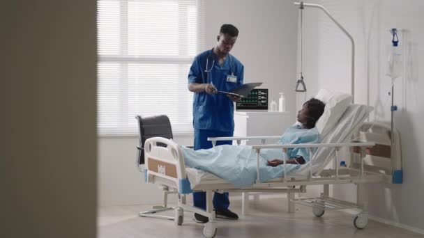 Mujer afroamericana descansando en cama de hospital después de la cirugía hablando con un joven enfermero. Retrato de enfermera afroamericana asistiendo a paciente enferma acostada en la cama — Vídeo de stock