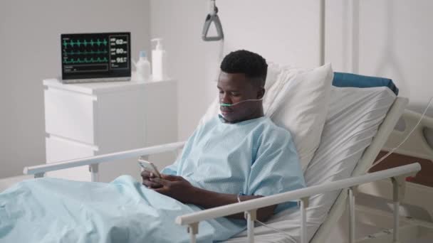 Afrikai férfi páciens okos telefonnal fekszik az ágyon. Afrikai beteg telefonon beszél a kórházi szobában — Stock videók