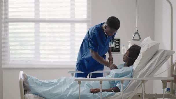 Mujer afroamericana descansando en cama de hospital después de la cirugía hablando con un joven enfermero. Retrato de enfermera afroamericana asistiendo a paciente enferma acostada en la cama — Vídeo de stock