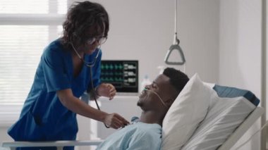 Siyahi bir kadın doktor, hastane yatağında yatan ve asit kaynağıyla bağlantısı olan siyah bir erkek hastayla konuşuyor.