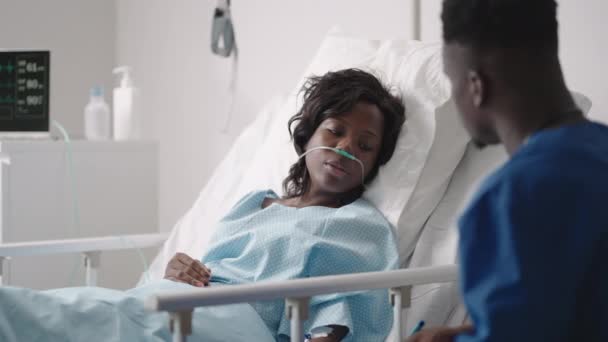 Mujer afroamericana descansando en cama de hospital después de la cirugía hablando con un joven enfermero. Retrato de enfermera afroamericana asistiendo a paciente enferma acostada en la cama — Vídeos de Stock