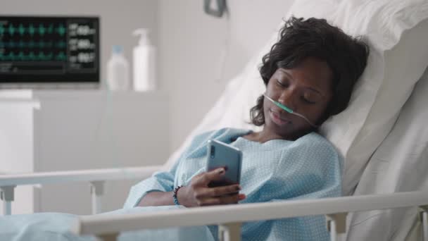 Paziente donna nera in ospedale con soluzione salina Pompa per infusione volumetrica utilizzando il telefono cellulare sul divano dell'esame. Donne africane sdraiate nel letto d'ospedale con smart phone mentre si trovano in ospedale. — Video Stock
