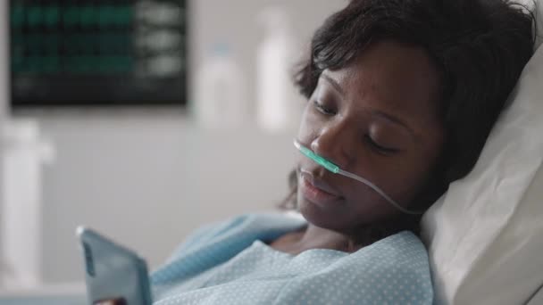 Czarna kobieta Pacjentka w szpitalu z solą fizjologiczną Pompa infuzyjna wykorzystująca telefon komórkowy na kanapie do badań. Afrykańskie kobiety leżące w szpitalnym łóżku ze smartfonem podczas pobytu w szpitalu. — Wideo stockowe