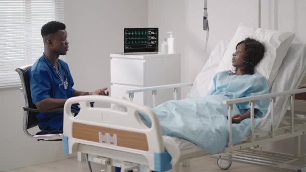 Afrikalı bir erkek doktor, hastane yatağında oksijen maskesi takmış bir hastayla röportaj yapıyor. Hastane yatağında yatan siyahi bir kadın doktora belirtileri anlatıyor. — Stok video