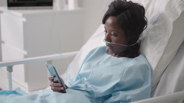 Черная женщина Пациент в больнице с раствором физраствора объемный инфузионный насос с помощью мобильного телефона на диване обследования. Африканские женщины лежат в больничной койке со смартфоном в больнице. — стоковое видео