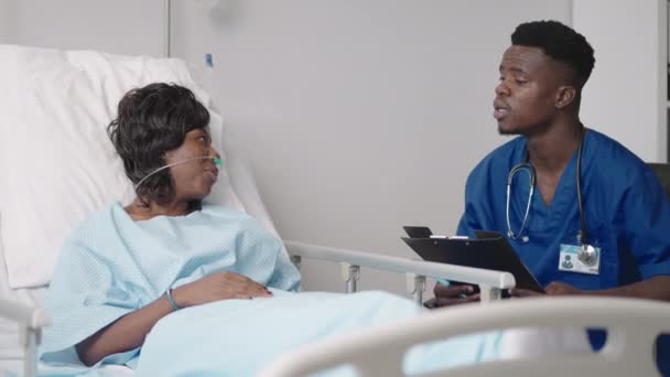 Afrykański lekarz przesłuchuje pacjenta leżącego w szpitalnym łóżku w masce tlenowej. Czarna kobieta leżąca w szpitalnym łóżku opisuje objawy lekarzowi. — Wideo stockowe