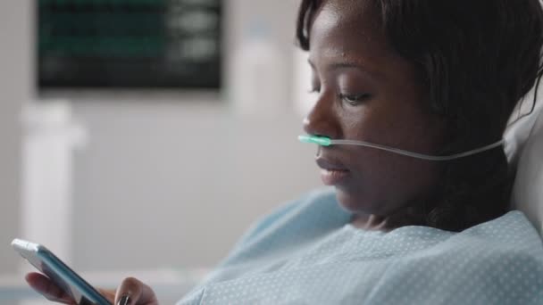 Paziente donna nera in ospedale con soluzione salina Pompa per infusione volumetrica utilizzando il telefono cellulare sul divano dell'esame. Donne africane sdraiate nel letto d'ospedale con smart phone mentre si trovano in ospedale. — Video Stock