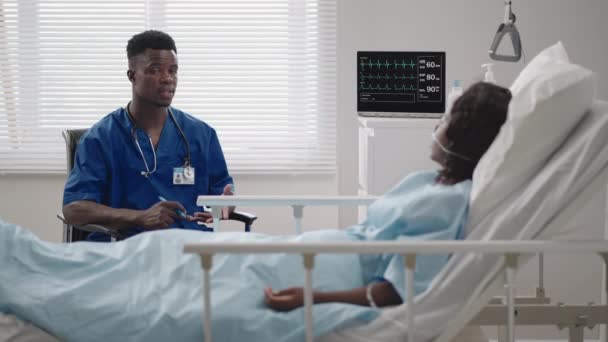 Un medico nero di sesso maschile sta parlando con una paziente nera distesa su un letto d'ospedale e collegata ad una scorta di acido. Il medico curante discute con il paziente in ospedale il trattamento — Video Stock
