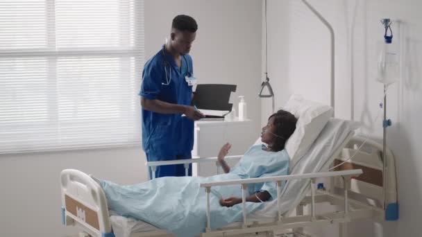 Un médico negro está hablando con una paciente negra acostada en una cama de hospital y conectada a un suministro de ácido. El médico que atiende discute con el paciente en el hospital el tratamiento — Vídeos de Stock