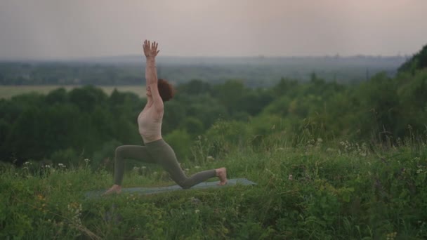 Una mujer joven hace ejercicios de yoga de pie en la cima de una montaña forestal sobre el fondo de un río y un paisaje forestal. Equilibrio de cuerpo y espíritu, relajación y meditación — Vídeos de Stock