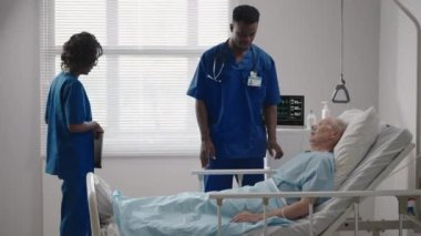 Yatakta yatan kıdemli hasta hastane odasında iki Afrikalı Amerikalı doktorla konuşuyor. Sağlık ve sigorta konsepti. Doktor yaşlı hastayı hastane yatağında rahatlatıyor ya da sağlık danışmanlığı yapıyor..