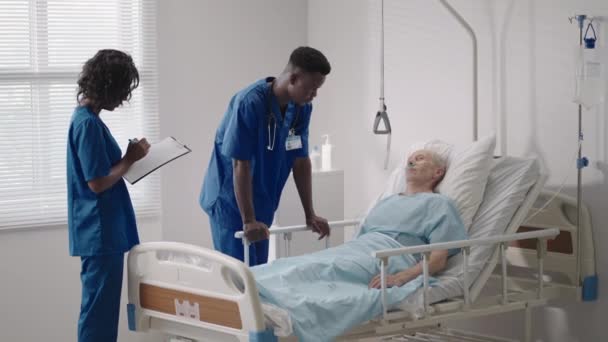 침대에 있는 선임 환자 가 병실에 있는 2 명의 아프리카 계 미국인 의사와 이야기하고 있습니다. 건강 관리와 보험 컨셉이죠. 병상에 있는 연로 한 환자를 위로 하거나 건강 진단을 하는 의사. — 비디오