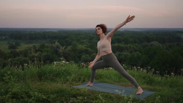 A young woman does yoga exercises standing on the top of a forest mountain against the background of a river and a forest landscape. Balance of body and spirit, relaxation and meditation — Stock Video