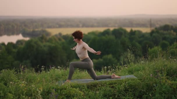 Una mujer joven hace ejercicios de yoga de pie en la cima de una montaña forestal sobre el fondo de un río y un paisaje forestal. Equilibrio de cuerpo y espíritu, relajación y meditación — Vídeos de Stock