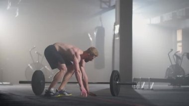 Ağır çekimde gömleksiz genç bir adam spor salonundaki bir halterin üzerinden atlıyor.