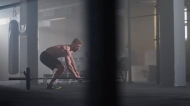 Ağır çekimde gömleksiz genç bir adam spor salonundaki bir halterin üzerinden atlıyor.