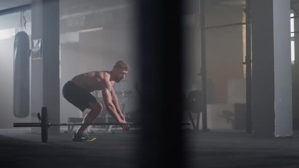 Een jongeman zonder shirt in slow motion springt een boertje over een lange halter in de sportschool in backlight — Stockvideo