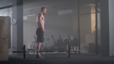 Ağır çekimde gömleksiz genç bir adam spor salonundaki bir halterin üzerinden atlıyor.