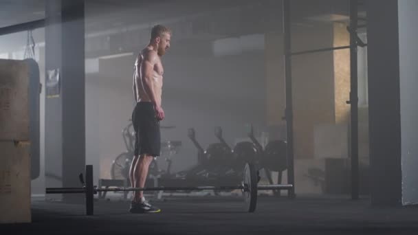 Молодой человек без рубашки в замедленной съемке прыгает отрыжкой через штангу в спортзале на заднем плане — стоковое видео