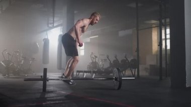 Ağır çekimde gömleksiz genç bir adam spor salonundaki bir halterin üzerinden atlıyor.