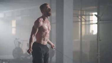 Tanınmış kasları olan genç bir atletik spor salonunda ağır çekimde ipe atlıyor.
