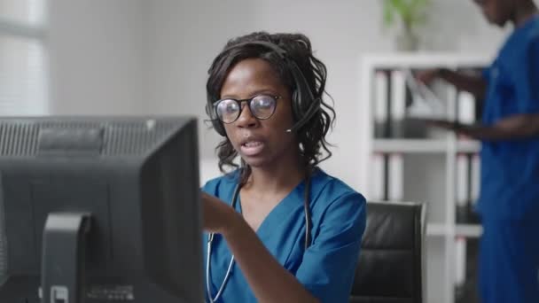 Una doctora negra que usa auriculares se sienta en una mesa con una computadora y recibe llamadas de pacientes mira sus registros médicos y los ingresa en el programa de la clínica. — Vídeo de stock