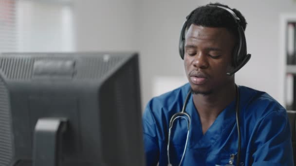 Afroamerikanische männliche Arzt oder Krankenschwester mit Headset und Computer arbeiten im Krankenhaus. Junge professionelle Therapeut Arzt Beratung Kunden mit Fernkommunikation sprechen auf Webcam — Stockvideo